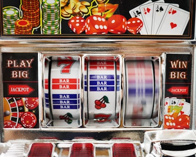 A close-up view of a slot machine with vibrant colors and various gambling symbols such as dice, coins, and playing cards. The reels display a combination of symbols, including bars, cherries, and the number seven. The chrome exterior and colorful details convey the theme of casino and gambling.