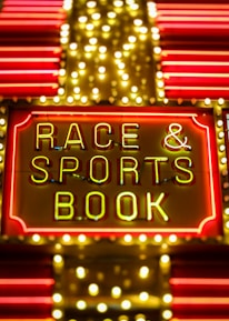 Bright neon lights on a marquee sign display the words 'RACE & SPORTS BOOK'. The sign is surrounded by rows of yellow and white lights against a red background, evoking a vibrant, energetic atmosphere typically associated with casinos or entertainment venues.