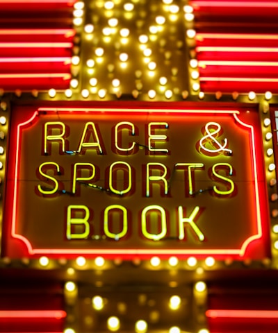 Bright neon lights on a marquee sign display the words 'RACE & SPORTS BOOK'. The sign is surrounded by rows of yellow and white lights against a red background, evoking a vibrant, energetic atmosphere typically associated with casinos or entertainment venues.
