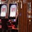 A pair of casino slot machines, each with detailed screens displaying various game outcomes and jackpot amounts. The chairs in front of the slot machines are dark and cushioned for comfort. The background features a hanging curtain made of golden ropes and other casino decorations are visible.