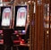 A pair of casino slot machines, each with detailed screens displaying various game outcomes and jackpot amounts. The chairs in front of the slot machines are dark and cushioned for comfort. The background features a hanging curtain made of golden ropes and other casino decorations are visible.