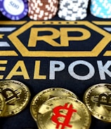Stacks of poker chips in various colors are arranged in the background, while in the foreground, there are numerous golden coins featuring the Bitcoin symbol. The text 'REALPOKER' is prominently displayed in gold and silver lettering on a dark background.