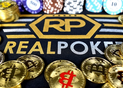 Stacks of poker chips in various colors are arranged in the background, while in the foreground, there are numerous golden coins featuring the Bitcoin symbol. The text 'REALPOKER' is prominently displayed in gold and silver lettering on a dark background.