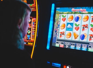 A person is closely observing a digital slot machine with colorful fruit symbols, bars, and a bell. The slot machine screen displays symbols like oranges, plums, cherries, and clouds. The background is dimly lit, creating a focused and somewhat intense atmosphere.