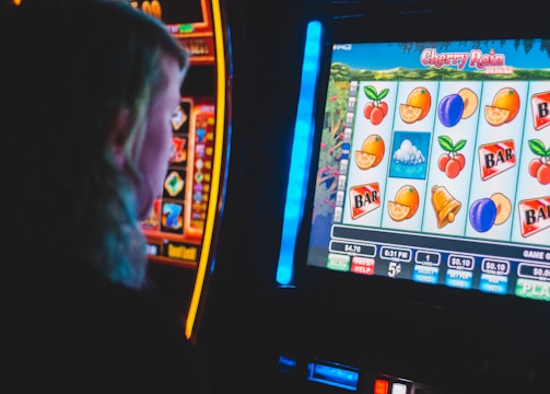 A person is closely observing a digital slot machine with colorful fruit symbols, bars, and a bell. The slot machine screen displays symbols like oranges, plums, cherries, and clouds. The background is dimly lit, creating a focused and somewhat intense atmosphere.