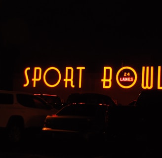 The image depicts a brightly lit neon sign that reads 'SPORT BOWL' with an additional sign indicating '24 LANES'. The scene appears to be taken at night, with several vehicles parked in front of the establishment.
