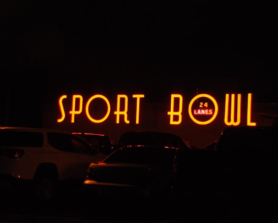 The image depicts a brightly lit neon sign that reads 'SPORT BOWL' with an additional sign indicating '24 LANES'. The scene appears to be taken at night, with several vehicles parked in front of the establishment.