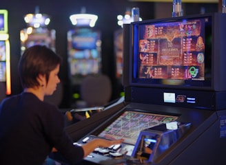 A person is seated at a slot machine console in a casino, focusing intently on the game displayed on the screen. The background shows several other illuminated gaming machines, adding a vibrant and colorful atmosphere to the scene.