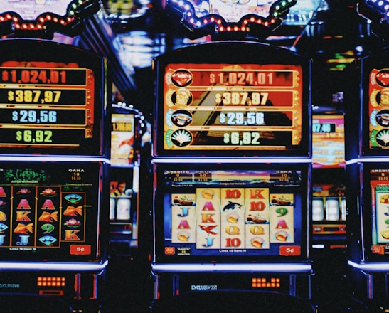 A row of colorful slot machines lit with bright neon lights, showcasing various graphical elements on the screens, indicating different games and jackpots. The atmosphere suggests a lively casino setting, bustling with excitement.