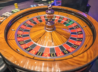 A roulette wheel with alternating red and black numbered pockets on a wooden platform. The wheel contains a small white ball resting in one of the pockets, indicating gameplay. Surrounding the wheel are stacks of colorful poker chips, suggesting a casino environment.