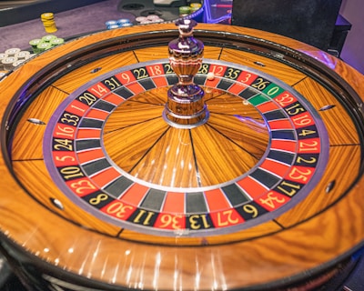 A roulette wheel with alternating red and black numbered pockets on a wooden platform. The wheel contains a small white ball resting in one of the pockets, indicating gameplay. Surrounding the wheel are stacks of colorful poker chips, suggesting a casino environment.