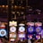 A lively casino scene featuring a row of bright, colorful slot machines with themes like Monopoly, Michael Jackson, and Lord of the Rings. The gaming machines display a variety of flashing lights and digital screens, creating an energetic and exciting atmosphere. Reflections of the lights are visible in glossy surfaces above the machines.
