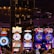 A lively casino scene featuring a row of bright, colorful slot machines with themes like Monopoly, Michael Jackson, and Lord of the Rings. The gaming machines display a variety of flashing lights and digital screens, creating an energetic and exciting atmosphere. Reflections of the lights are visible in glossy surfaces above the machines.