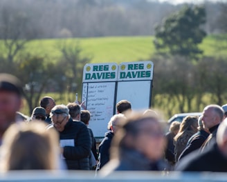 A group of people gathered outdoors with two large betting boards in the background labeled 'REG DAVIES'. The setting appears to be a countryside location with trees and fields visible in the distance. The crowd consists of individuals dressed in jackets and hats, engaging in conversation.
