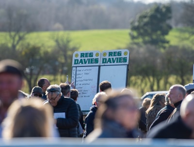 A group of people gathered outdoors with two large betting boards in the background labeled 'REG DAVIES'. The setting appears to be a countryside location with trees and fields visible in the distance. The crowd consists of individuals dressed in jackets and hats, engaging in conversation.