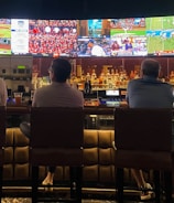 A group of people sit at a bar with their backs to the camera, watching multiple sports games displayed on large screens. The bar counter is lined with various bottles of liquor, and the atmosphere suggests a casual and relaxed social setting.