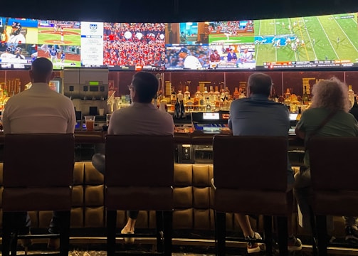 A group of people sit at a bar with their backs to the camera, watching multiple sports games displayed on large screens. The bar counter is lined with various bottles of liquor, and the atmosphere suggests a casual and relaxed social setting.