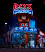 A vibrant, neon-lit facade of a gaming and sports establishment at night, featuring a large glove holding a baseball above the entrance. The building is adorned with colorful signs in both English and Japanese, with various advertisements and an illuminated sign that says 'ROX DOME'. Several people are walking towards the entrance, and bicycles are parked along the side.