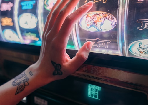 A hand with tattoos reaches towards a brightly lit slot machine, featuring various colorful symbols including a tiger. The image captures the moment of anticipation as the person engages with the machine during a game of chance.